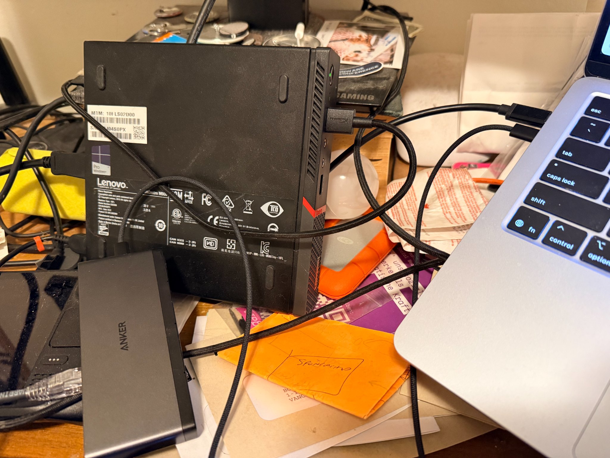 An extremely messy desk - a Lenovo m900 tiny is plugged into an Ethernet jack on a Mac dock into a Macbook Air and it only has displayport into a big monitor behind it. There is detritus on the desktop.