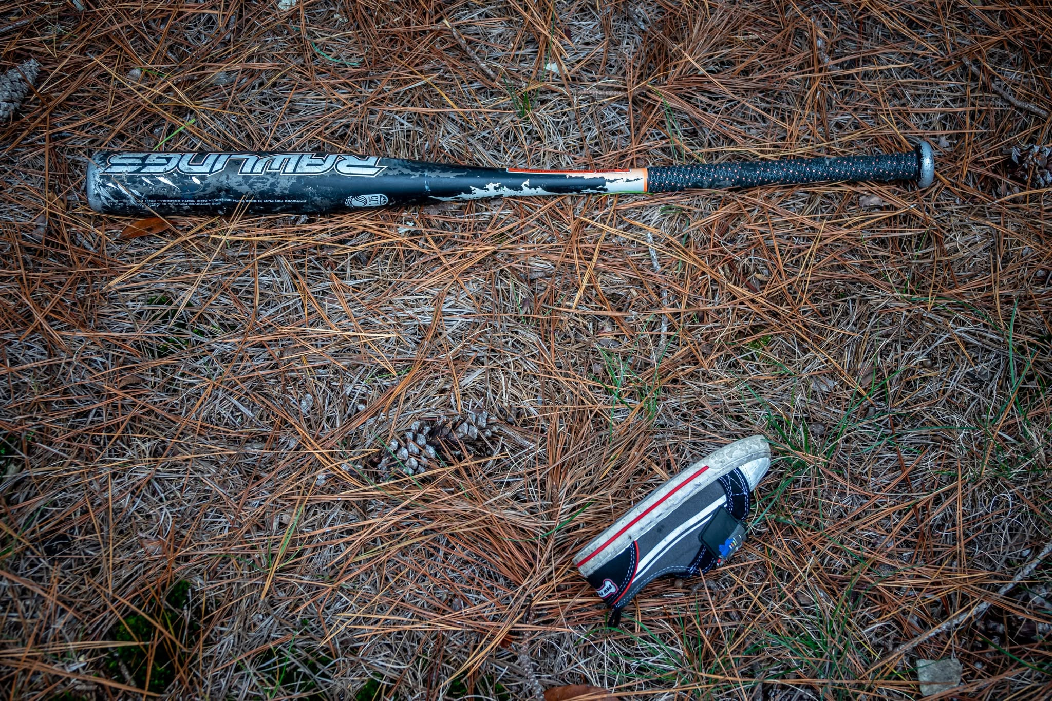 An overhead view shows a discarded black-and-silver Rawlings youth baseball bat and a small child's sneaker lying on a bed of dry pine needles. The scuffed metal bat is positioned horizontally across the upper portion of the frame, showing signs of wear and chipped paint. In the lower right corner, a single black-and-gray canvas shoe with a red stripe and white toe sits angled alongside a scattered pinecone.
