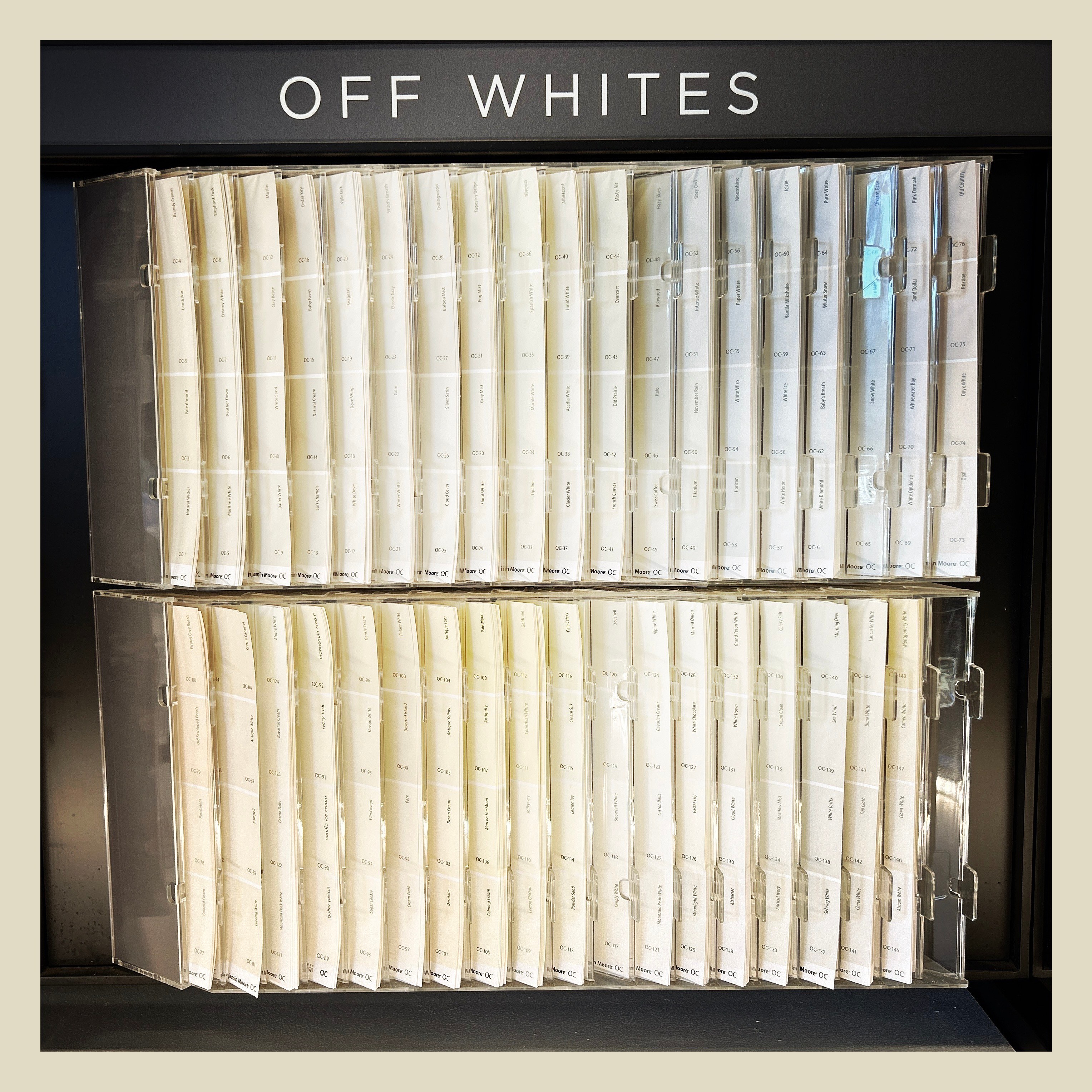 A retail paint display featuring a dark header labeled "OFF WHITES" in white capital letters is organized into two stacked acrylic racks. The transparent slots are filled with numerous vertical paint swatch strips showing subtle variations of white, cream, beige, and light gray tones. Each color card is neatly aligned in columns and labeled with individual paint names and reference codes.